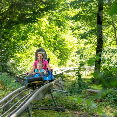 Alpine-Coaster-Golm in Montafon | © Golm Silvretta Lünersee Tourismus GmbH Bregenz, Stefan Kothner A woman and a child ride down through a forest on a summer roller coaster | © Golm Silvretta Lünersee Tourismus GmbH Bregenz, Stefan Kothner