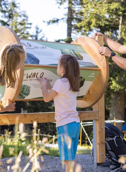 Golmis Forschungspfad at Erlebnisberg Golm | © Golm Silvretta Lünersee Tourismus GmbH Bregenz, Stefan Kothner A father with two children on Golmis Forschungspfad. They are turning at a rectangular information board with lots of information about the forest. | © Golm Silvretta Lünersee Tourismus GmbH Bregenz, Stefan Kothner
